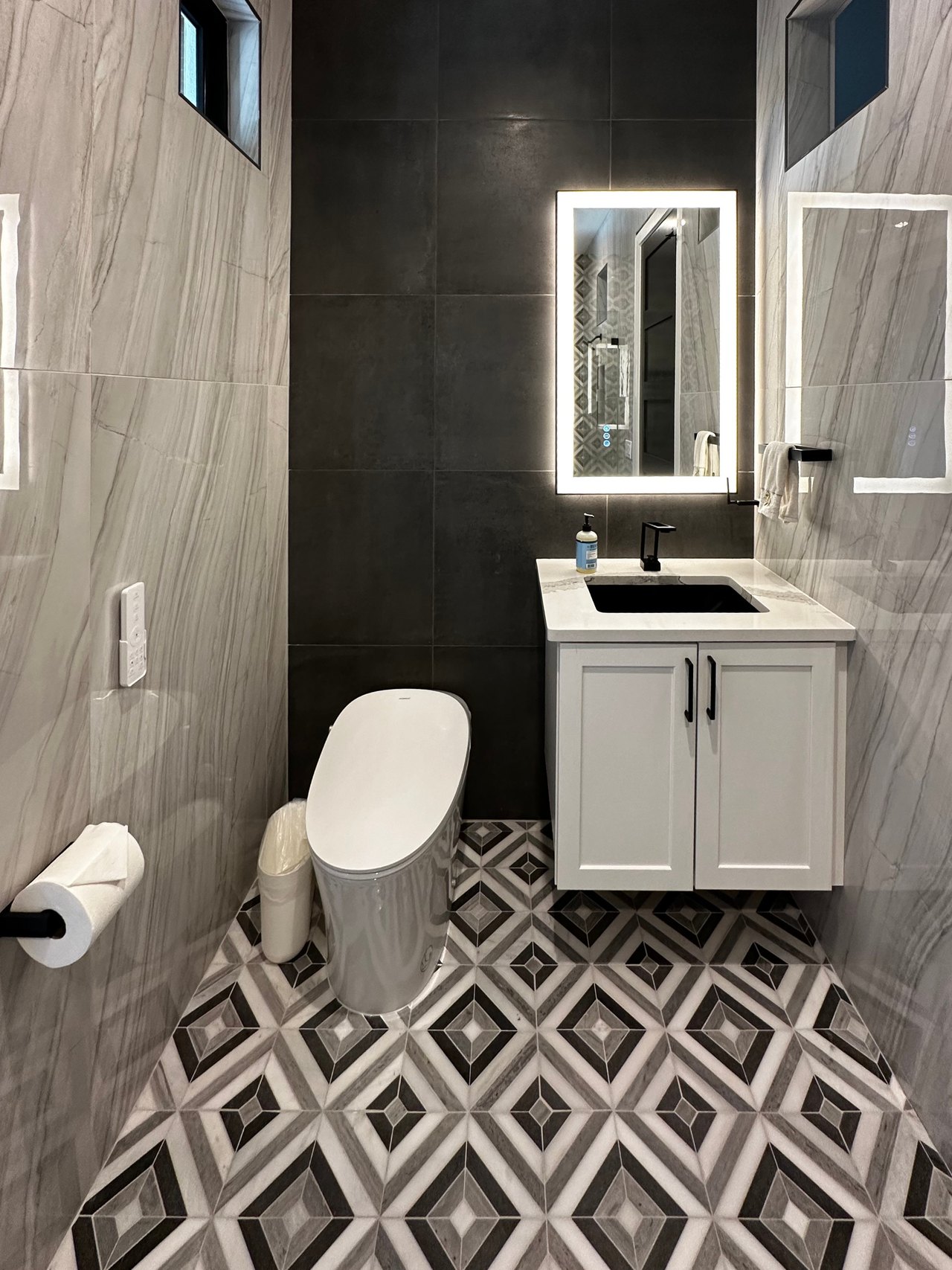 Bathroom with a floating white vanity, illuminated mirror, charcoal accent wall and geometric floor tile