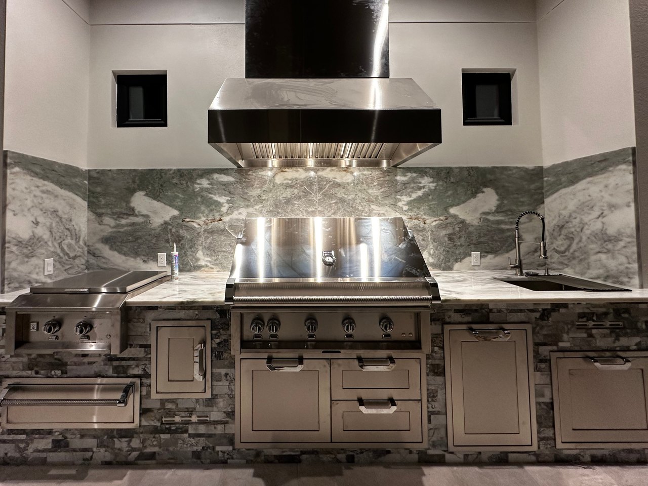 Outdoor kitchen with a stainless steel grill, vent hood, griddle and sink with a stone-clad base