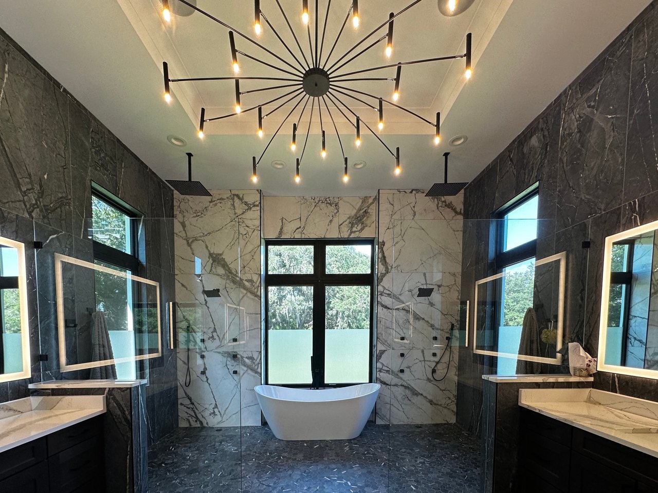 Primary bathroom with a centered freestanding tub, glass showers, dual vanities and a sculptural chandelier