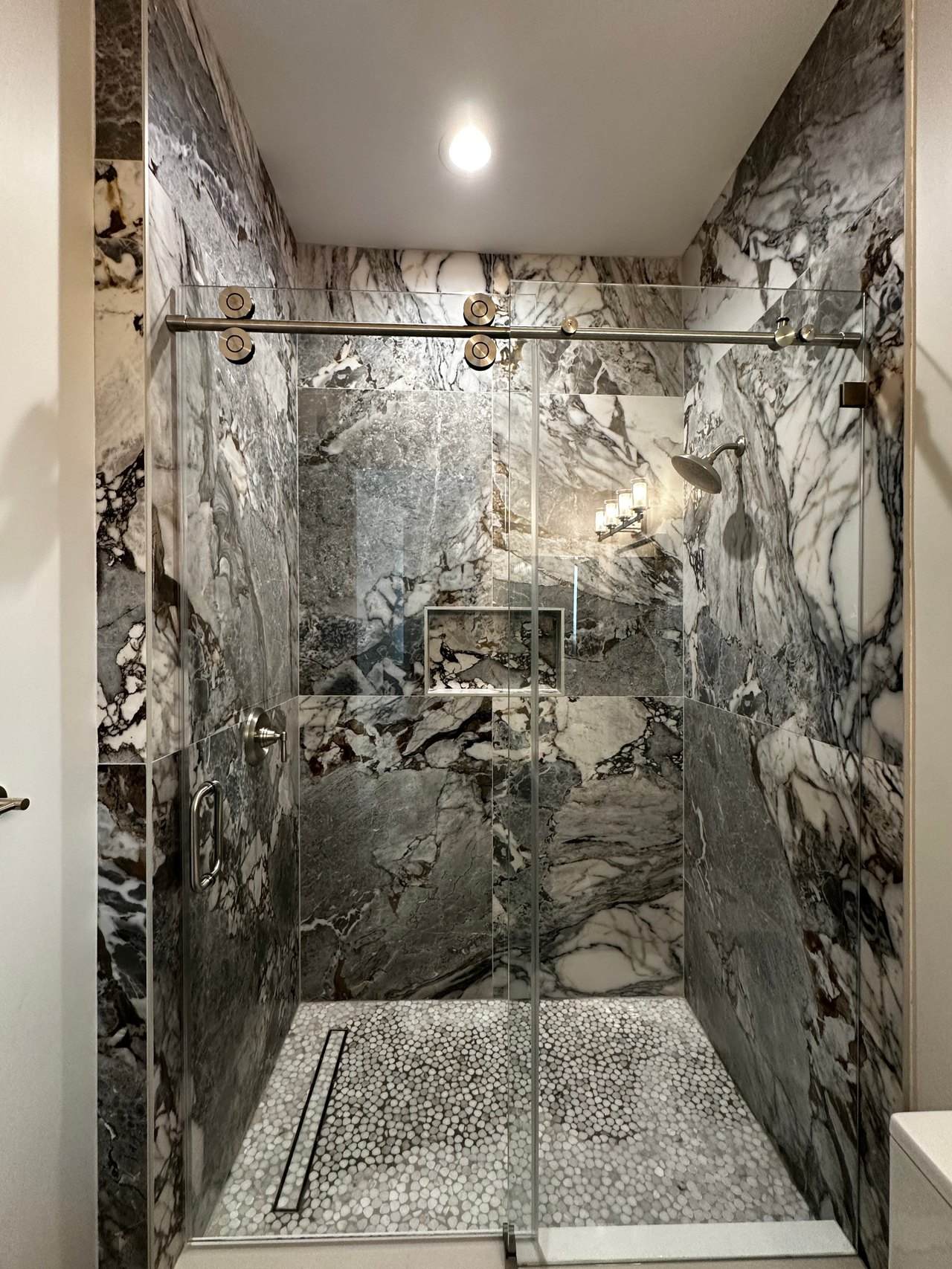 Walk-in shower with veined stone-look walls, a sliding glass door and a pebble tile floor